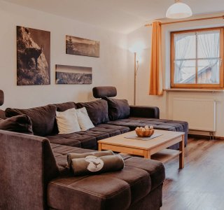 Ein Wohnzimmer mit einem großen Sofa und einem Tisch. Die Fenster lassen viel Licht herein. Es ist eine ruhige Atmosphäre.