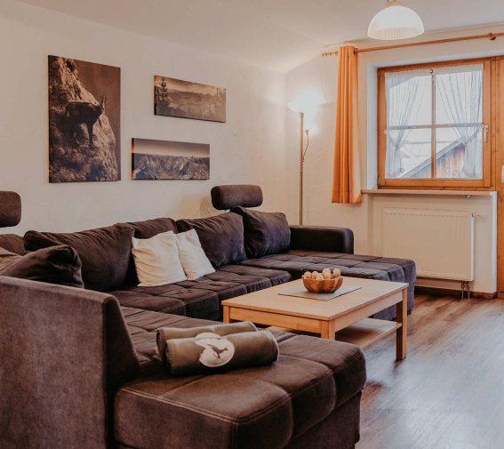 Ein Wohnzimmer mit einem großen Sofa und einem Tisch. Die Fenster lassen viel Licht herein. Es ist eine ruhige Atmosphäre.