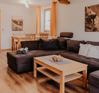 Im Raum sind eine große Couch, ein Tisch und ein Essbereich. Die Wände haben Bilder und das Licht ist hell.