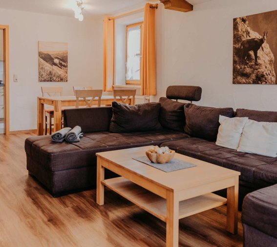 In einem Wohnzimmer mit einer Couch und einem Tisch Im Raum sind eine große Couch, ein Tisch und ein Essbereich. Die Wände haben Bilder und das Licht ist hell.