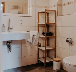 Badezimmer mit einfacher Einrichtung und Waschbecken In einem Badezimmer stehen ein Waschbecken und ein Regal. Ein Handtuch hängt am Waschbecken. Es ist hell und sauber.