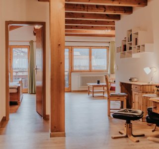 Ein heller Raum mit Holzdecken und Fenstern. Möbel stehen ordentlich, und es gibt einen Arbeitsplatz mit Stuhl.