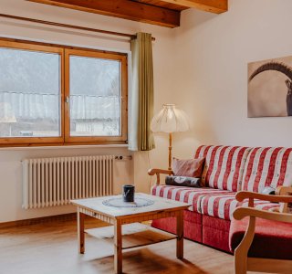 In einem Wohnbereich befindet sich ein rotes Sofa und ein Tisch. Es ist hell und ein Fenster zeigt die Berge draußen.