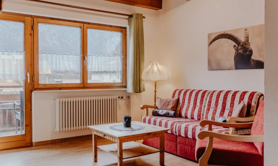 In einem Wohnbereich befindet sich ein rotes Sofa und ein Tisch. Es ist hell und ein Fenster zeigt die Berge draußen.