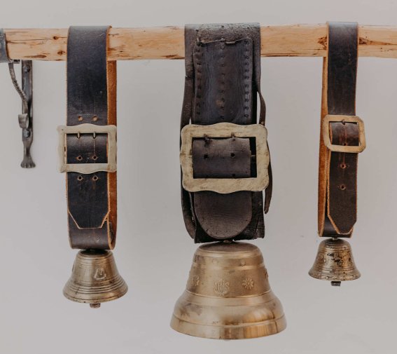 Drei Gürtel mit Glocken hängen an einem Holzstab. Der Platz ist hell und die Gürtel sind einfach angeordnet.