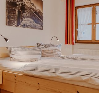 Der Raum zeigt ein Doppelbett mit gestreiften Bettlaken und einer Fensteransicht. Es gibt eine Holzkommode und Wandbilder.