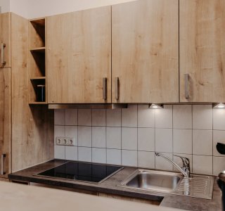 In der Küche sind Holzschränke, ein Wasserhahn und ein Herd zu sehen. Licht fällt auf die Arbeitsfläche und die Wand.