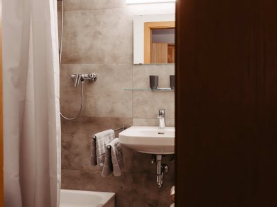 In einem hellen Badezimmer stehen ein Waschbecken und eine Dusche vor grauen Fliesen. Handtücher hängen bereit.