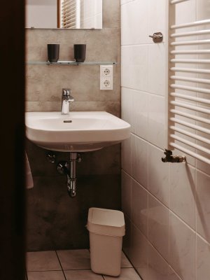 Das Badezimmer zeigt ein Waschbecken mit Armatur und einem Abfallkorb. Es hat helle Fliesen und klare Linien.