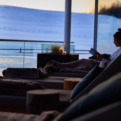 Ein Gast liest ein Buch im Ruheraum im Fasten-Hotel Allgäu, während die Sonne hinter den schneebedeckten Bergen versinkt.