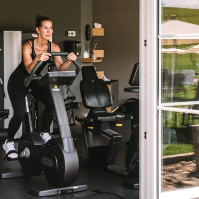 Eine Frau trainiert auf einem Fahrrad-Ergometer bei offenen Fenstern im Fitnessraum des Sporthotels.