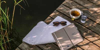 Auf dem Steg des Haubers Natursees liegen Handtuch, Sonnenbrille, Tee und ein Buch im Schatten des Schilfs.