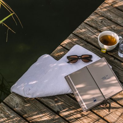 Auf dem Steg des Haubers Natursees liegen Handtuch, Sonnenbrille, Tee und ein Buch im Schatten des Schilfs.