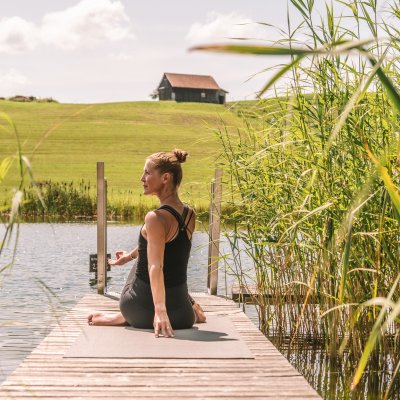 Eine Frau sitzt auf einer Yogamatte auf am Rand des Hotelsees und dreht ihren Oberkörper nach links.