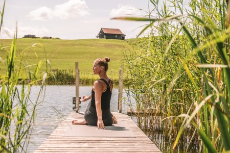 Eine Frau sitzt auf einer Yogamatte auf am Rand des Hotelsees und dreht ihren Oberkörper nach links.