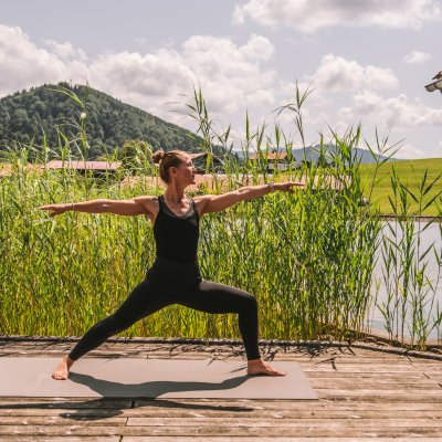 Vor dem Allgäuer Panorama praktiziert eine Gästin Yoga am Natursee des Wellnesshotels.