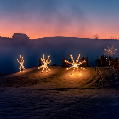 Mit Lichterketten verzierte Strohsterne stehen rund um das Wellnesshotel Haubers in der Dämmerung im Schnee.