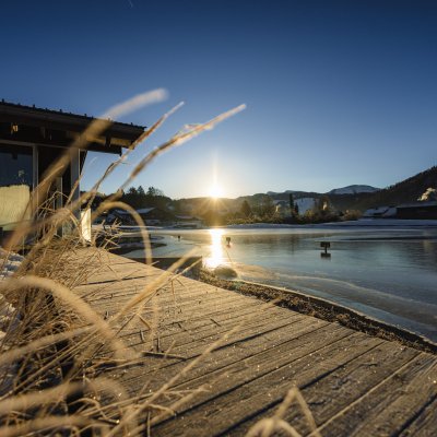 Über dem gefrorenen Natursee von Haubers Wellnessbereich geht die Morgensonne auf und lässt den Schnee tauen.
