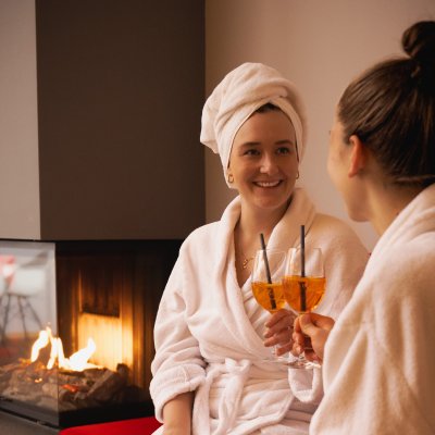 Zwei Freundinnen genießen ihren Drink im Bademantel in Haubers Spa am Kaminfeuer.