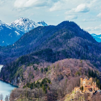 Schloss Hohenschwangau vor Alpenpanorama. Hinter dem alten Schloss Hohenschwangau sind der Alpsee und schneebedeckten Gipfel in der Ferne sichtbar.