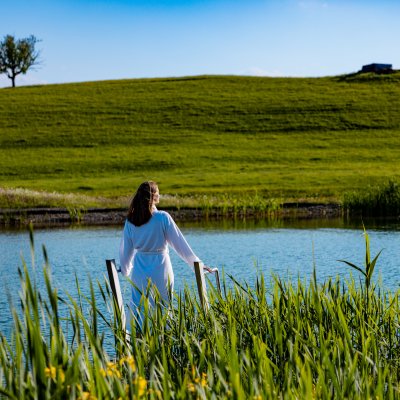 Am Einstieg zum Badesee vor Haubers Naturresort steht eine Frau im Bademantel und genießt den Ausblick.