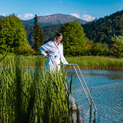 Eine Frau im Bademantel steht mit geschlossenen Augen an Haubers Natursee und lässt sich die Sonne ins Gesicht scheinen.