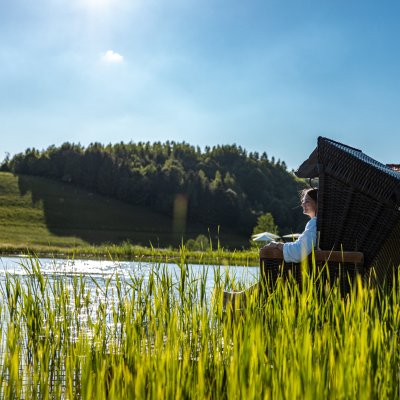 Eine Frau sitzt entspannt im Strandkorb und blickt über den Natursee, der in der Sommersonne funkelt.
