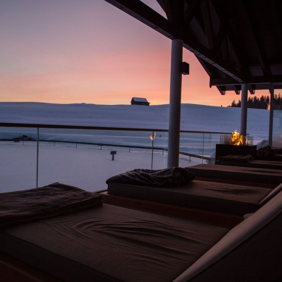 Das Allgäuer Abendrot taucht die Ruheliegen am verschneiten Natursee von Haubers Wellnesshotel in warmes Licht.