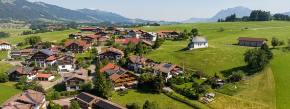 Ein Blick auf Dörfer mit Häusern in einer grünen Landschaft. Berge sind im Hintergrund sichtbar. Es ist sonnig und klar.