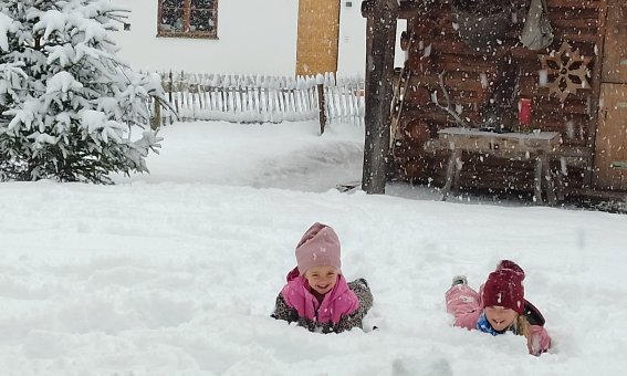 Zwei Kinder liegen im Schnee und lachen. Es schneit stark und der Garten ist weiß bedeckt.