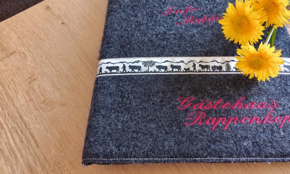 Eine Menükarte liegt auf einem Tisch. Auf der Karte steht der Name des Gastehauses und es sind Blumen dekoriert.