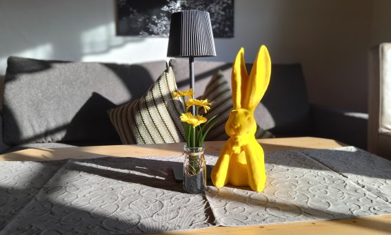 Auf einem Tisch steht ein gelber Hase neben einer Vase mit Blumen und einer Lampe. Die Sonne scheint durch das Fenster.