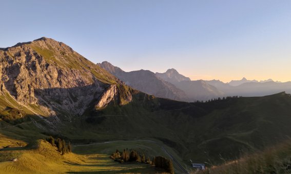 Die Sonne senkt sich hinter den Bergen und wirft Schatten auf das Tal. Die Landschaft ist ruhig und einladend.