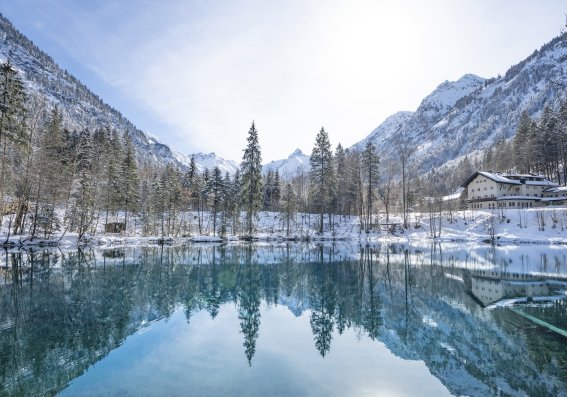 Ein ruhiger Bergsee spiegelt die schneebedeckten Berge und Wälder in der Wintersonne wider.