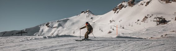 Ein Skifahrer fährt die verschneite Piste in den Bergen hinunter. Der Himmel ist klar und blau, die Sonne scheint.