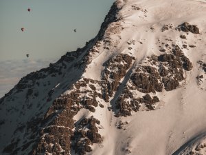 Mehrere Heißluftballons schweben über einem schneebedeckten Berggipfel an einem klaren, sonnigen Tag.