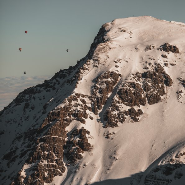 Mehrere Heißluftballons schweben über einem schneebedeckten Berggipfel an einem klaren, sonnigen Tag.