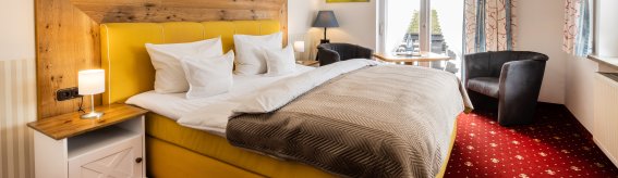 das Zimmer hat ein großes, gelbes Bett und einen Blick auf einen Balkon. Es gibt einen Stuhl und Bilder an der Wand.