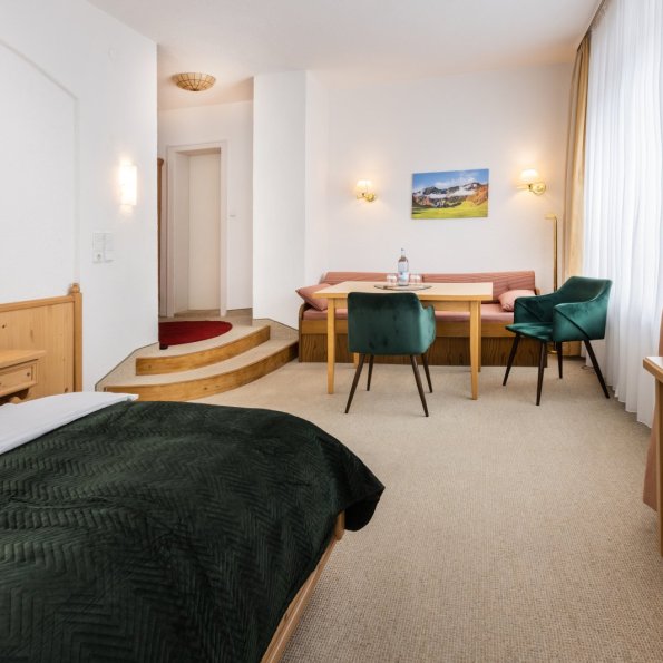 Das Zimmer zeigt ein Bett, einen Tisch und einen Fernseher. Es gibt viel Licht und Platz. Ein Bild hängt an der Wand.