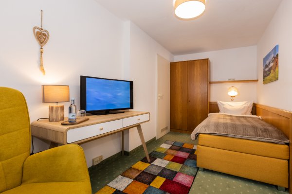 In einem kleinen Raum steht ein Bett, ein Fernseher und ein Sessel. Der Boden hat einen bunten Teppich.