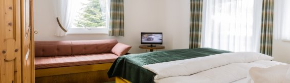 In einem Raum stehen ein Bett und eine Couch. Ein Fernseher ist gegenüber. Die Fenster sind hell und offen.