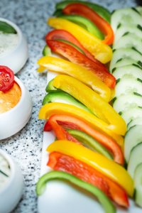 Auf einem weißen Teller liegen frisches Gemüse und Dips. Es gibt Paprika, Gurken und Tomaten sowie zwei Schalen mit Dips.