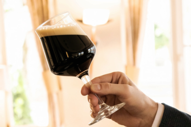 Eine Hand hält eine Biertulpe, gefüllt mit dunklem Craftbier in die Höhe.
