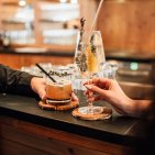 Drinks und Genuss an der Kaminbar im Hotel Oberstdorf Zwei Gäste stoßen mit Cocktails an der Kaminbar des Hotel Oberstdorf an – entspannte Genussmomente in gemütlicher Atmosphäre.