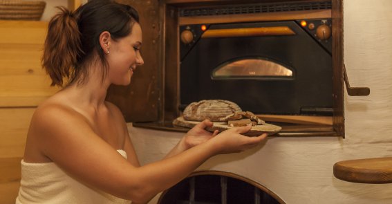 Eine Frau schiebt ein Brot in den Ofen in der Brotsauna im Hotel Oberstdorf Im Brotbad im Hotel Oberstdorf wird jeden Tag frisches Allgäuer Brot gebacken, das Sie an der Wellnessbar probieren dürfen. Die Hitze des Ofens erwärmt den ganzen Raum auf angenehme Biosauna-Temperatur.