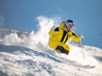 Ein Skifahrer fährt schnell den Hang hinunter. Schnee spritzt um ihn herum. Der Himmel ist blau und klar.