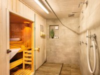 Eine Sauna steht in einem Wellnessbereich. Ein Duschbereich befindet sich daneben. Licht vom Holz sitzt in der Sauna.