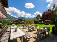Das Café zeigt eine Terrasse mit Tischen und Stühlen. Im Hintergrund sind Berge und Bäume sichtbar.