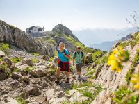 Zwei Personen wandern auf einem steinigen Weg in den Alpen. Sie tragen Rucksäcke und sehen nach oben.