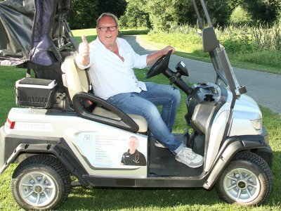Ein Mann fährt auf einem Golfplatz mit einem Golfcart und zeigt mit Daumen nach oben. Sonne scheint, entspannte Atmosphäre.